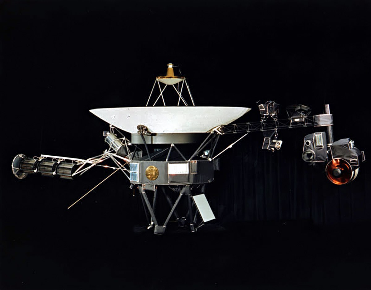 The Voyager 1 spacecraft, a complex assembly of scientific instruments, antennas, and power sources, set against a black background. Its large dish antenna is prominent in the center.