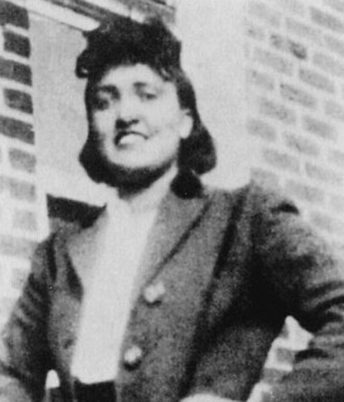 A black and white portrait of Henrietta Lacks, showing her smiling slightly and wearing a dark jacket with a light-colored collar. A brick wall is visible in the background.