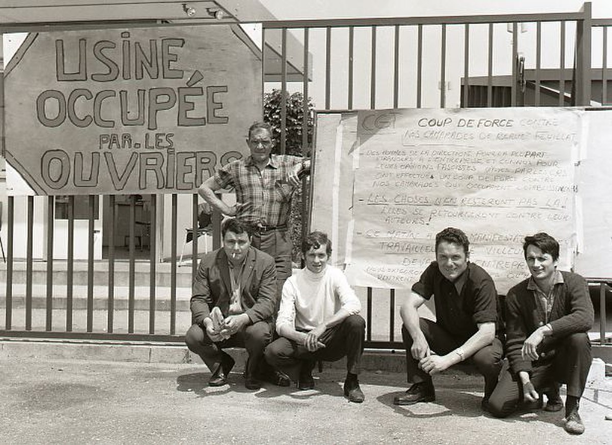 French workers pose in front of a factory entrance with a large sign reading "USINE OCCUPEE PAR LES OUVRIERS" (Factory Occupied by the Workers) during the May 1968 strikes. Another sign behind them lists demands, and the photo was taken in June 1968.