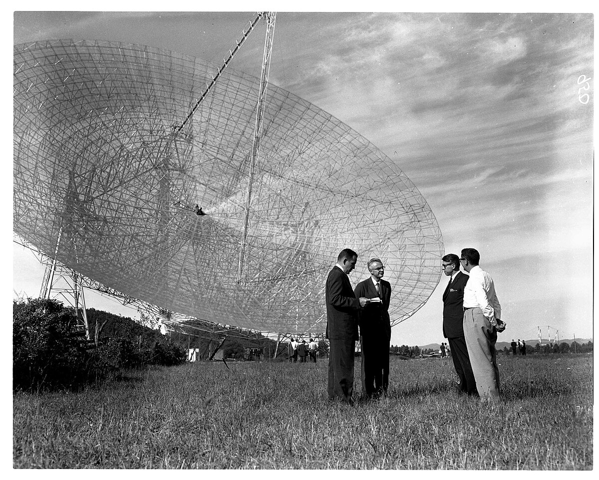 Frank Drake, second from left, stands with three other men in front of the completed 300-foot radio telescope. The large dish antenna dominates the background against a cloudy sky.
