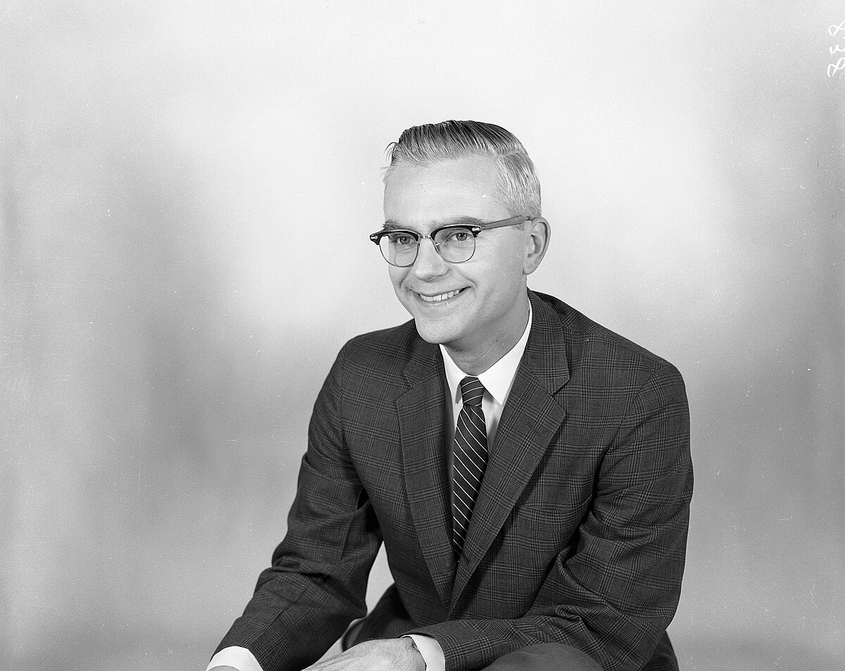 Portrait of Frank Drake in the 1960s, wearing glasses and a suit with a striped tie. He is smiling and looking to his right.