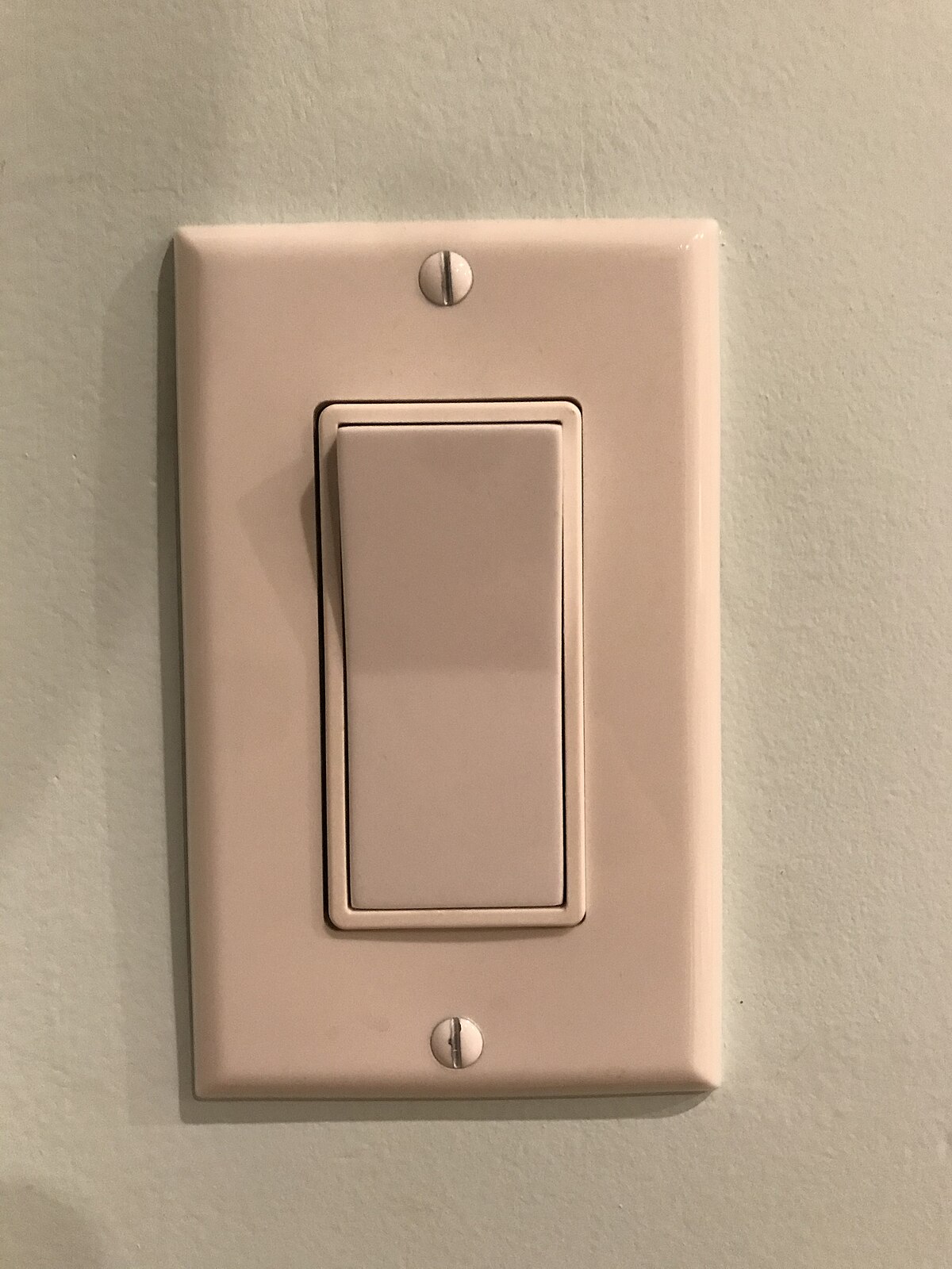 A close-up photograph of a standard rocker light switch on a light green wall. The switch is in the neutral, or off, position.