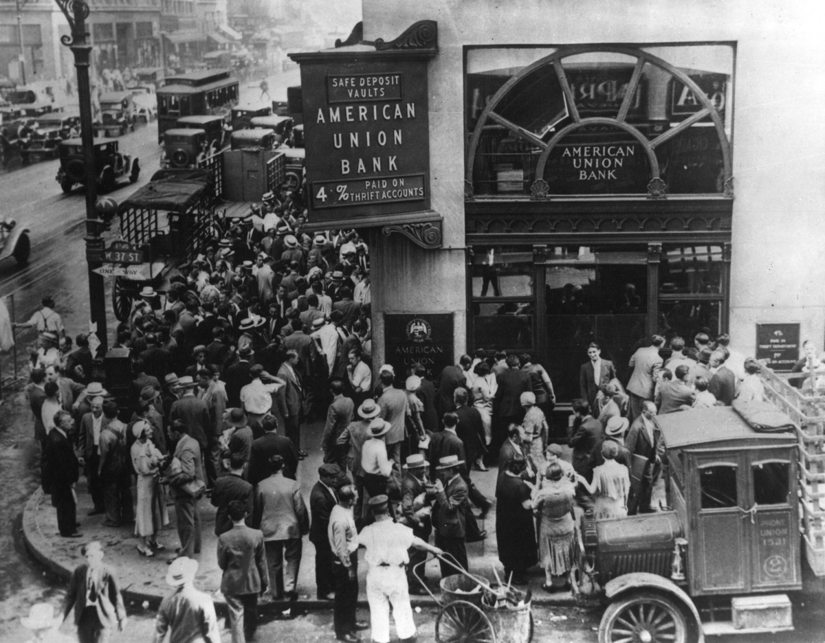 A large crowd of people gathered outside the American Union Bank in New York during a bank run early in the Great Depression. A sign on the building advertises "SAFE DEPOSIT VAULTS" and "4% PAID ON ALL ACCOUNTS."