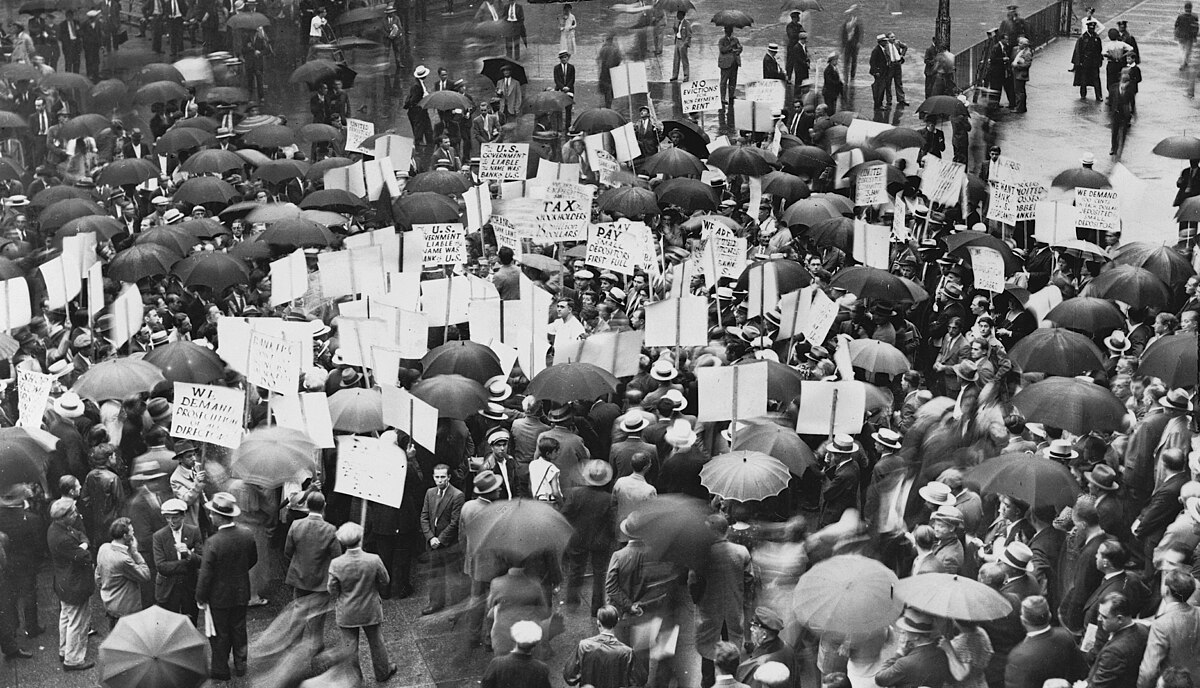 A large crowd of people, many holding umbrellas and protest signs, gathered outside the Bank of United States in New York in 1931 after its failure. Visible signs include "TAX PAYERS" and "WE WANT OUR MONEY."