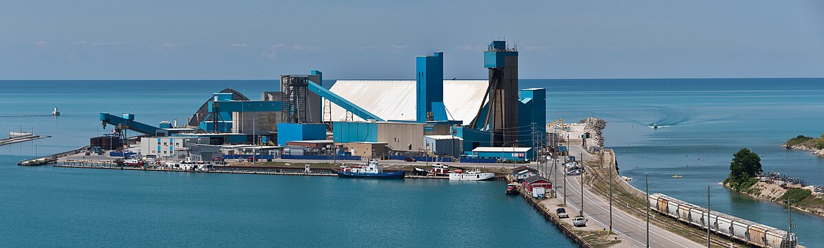 A panoramic view of the Sifto Canada salt mine and processing plant located at the harbor in Goderich, Ontario, Canada. The industrial complex features large blue and white buildings, conveyor belts, and ships docked in the blue water.