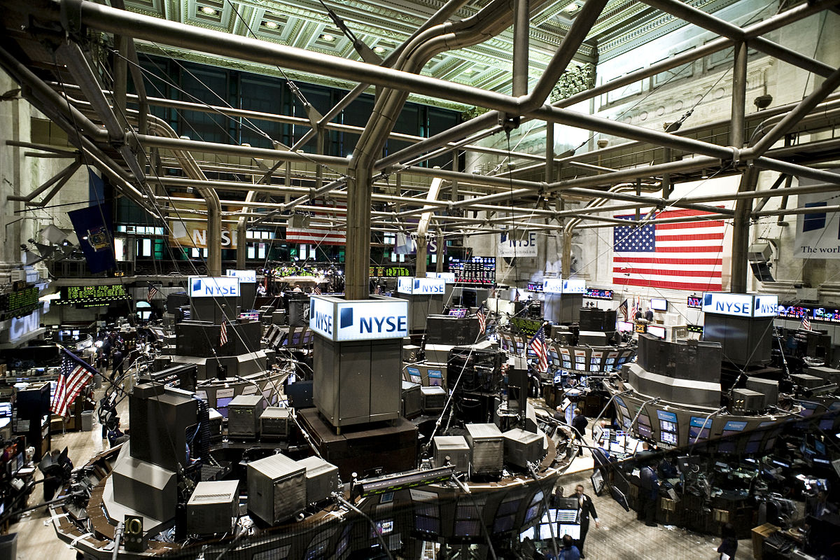The trading floor of the New York Stock Exchange in 2009, showing a busy scene with numerous traders and multiple trading posts equipped with screens. American flags are visible hanging from the ceiling.
