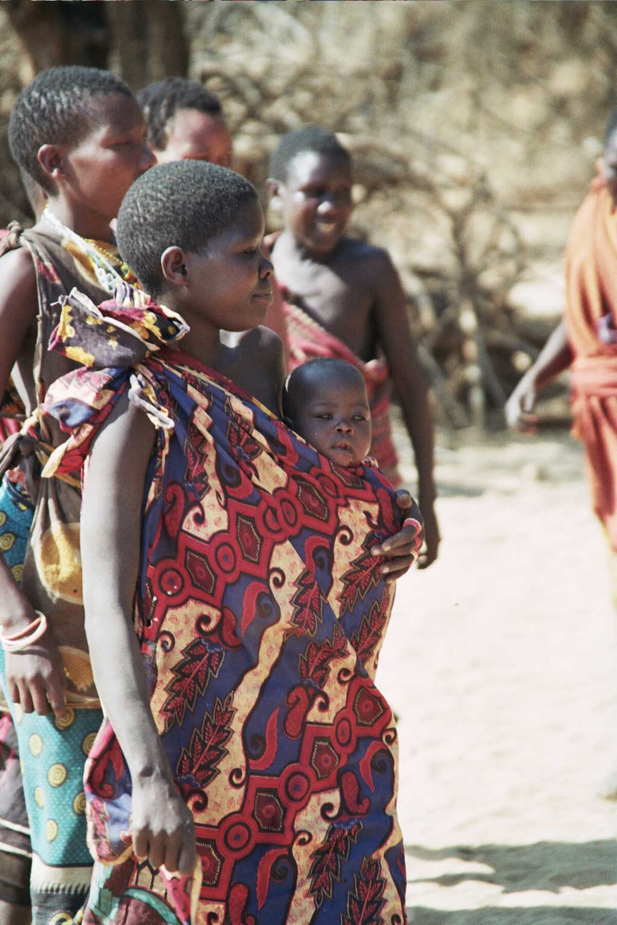 A Hadza woman carrying a baby in a colorful cloth sling, with other Hadza people in the background. The image captures a moment of daily life among the Hadza people, an indigenous ethnic group in Tanzania.