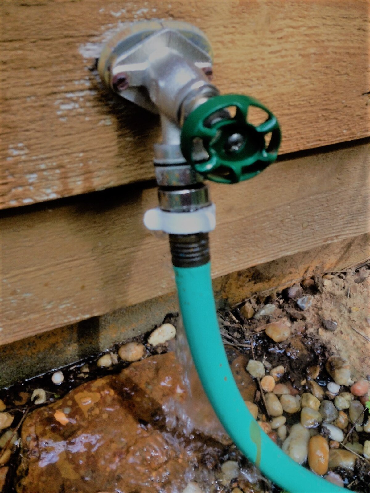 A leaking garden hose bib is shown attached to a wooden wall, with water visibly dripping from the connection to a turquoise garden hose. The green handle of the spigot is partially visible, and water is pooling on the rocky ground below.