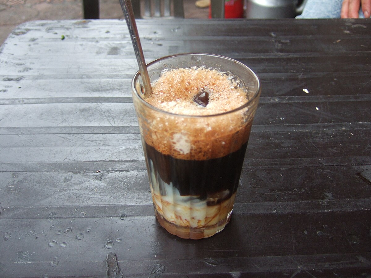 A glass of Vietnamese coffee (Cà Phê Sữa Đá) served with condensed milk, showing distinct layers of dark coffee, ice, and white condensed milk at the bottom. A spoon is in the glass, and it sits on a dark, textured surface.